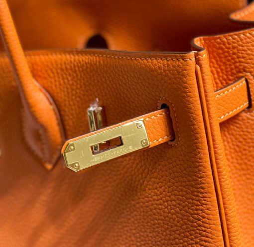 HERMÈS Birkin. Exquisite Craftsmanship and Timeless Elegance: Discover the Allure of the HERMES Birkin Bag Large Leather Top Handle. Designed with the actress Jane Birkin in mind, the Birkin bag is a contemporary development of the Kelly bag. Created based on Jane's suggestions, it was architected to be more significant to fit modern women's fast-paced life, with several minor yet outstanding adaptations, such as the elongated top handles. The French Birkin 30 bag is a classic touch of elegance to your look! The accessory features a trapeze body, a pebbled leather texture, a hanging key fob, gold-tone hardware, round top handles, a padlock fastening detail, a foldover top with twist-lock closure, a front center logo stamp, an internal zipped pocket, and purse feet.