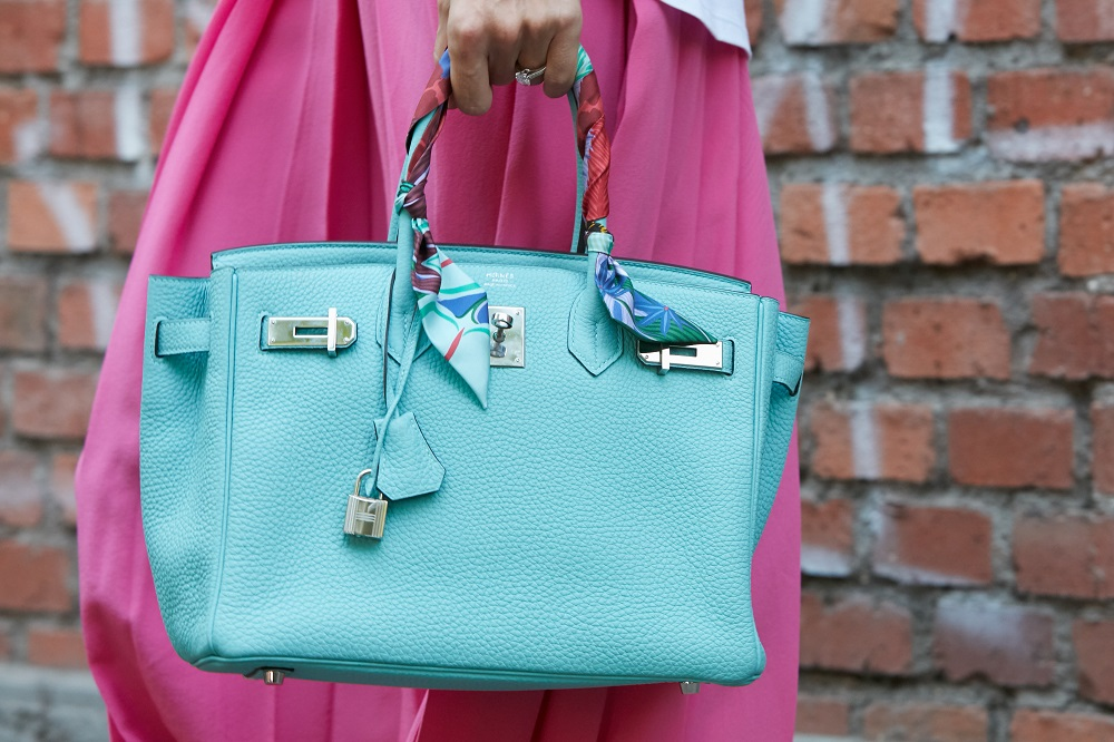 A vibrant turquoise Birkin with twilly-wrapped handles adds a pop of color against a brick wall backdrop.